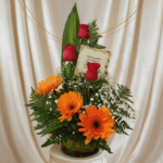 Arreglo floral en jarrón de vidrio con gerberas naranjas, rosas rojas, follaje verde y tarjeta sobre fondo de cortina beige