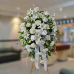 Corona fúnebre de rosas y lirios blancos con lazo blanco sobre caballete de madera en interior