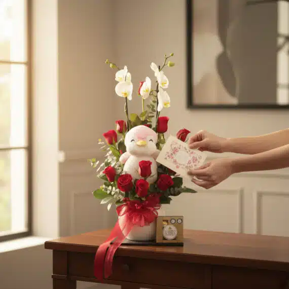 Manos delicadas colocando una tarjeta en el Arreglo ALICE con Orquídea Majestuosa, Rosas Rojas, peluche de pingüino y chocola