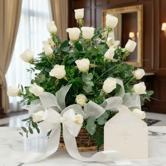 Arreglo floral blanco puro con rosas y follaje verde en una cesta de mimbre, decorado con una cinta de tela satinada blanca,