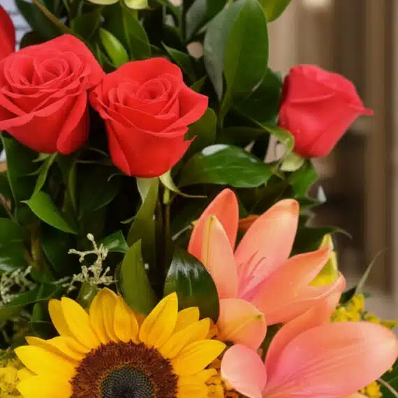 Primer plano extremo de un arreglo floral con rosas rojas, girasol, lirios naranjas y hojas verdes, destacando texturas y got