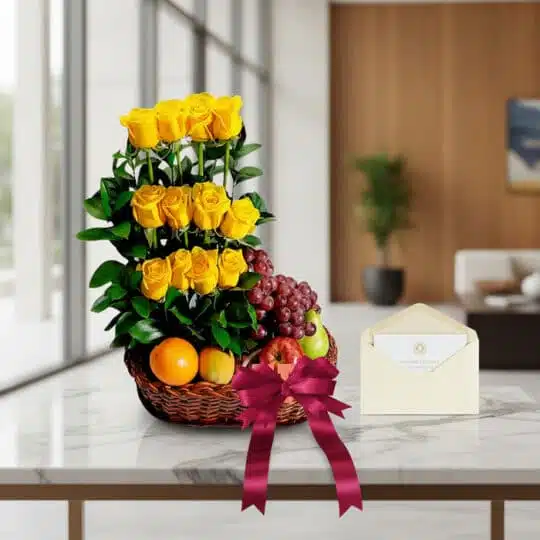 Arreglo floral con rosas amarillas y frutas frescas en cesta de mimbre, con tarjeta de regalo y cinta de satén, sobre mesa de