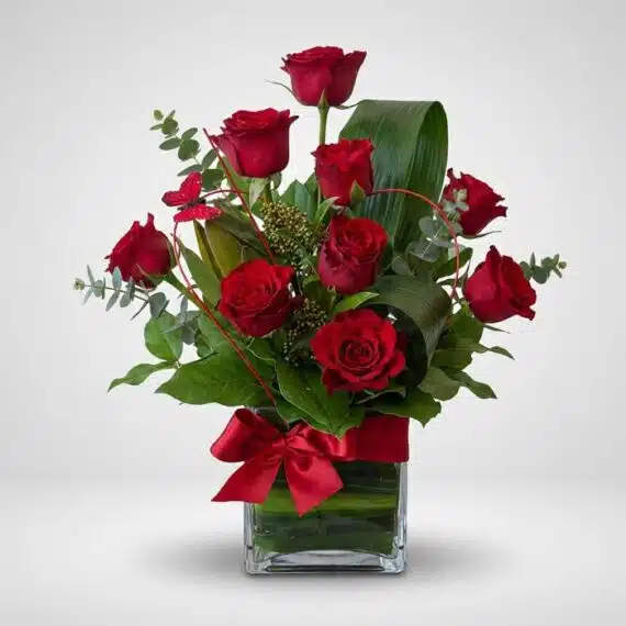 Arreglo Floral ROSETTA: Seis Rosas Rojas en Elegante Jarrón Arreglo floral ROSETTA con seis rosas rojas frescas, follaje verde y un lazo rojo en un jarrón de cristal, ideal para regalar