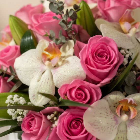 Primer plano extremo de un arreglo floral con rosas rosadas, orquídeas blancas con gotas de rocío, y follaje verde, destacand
