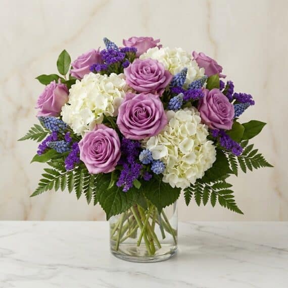 Arreglo Floral VITTORIA: Rosas Lilas y Hortensias para Regalar Arreglo floral VITTORIA con rosas lilas, hortensias blancas y muscari azul en un jarrón de cristal, sobre una mesa de mármol.