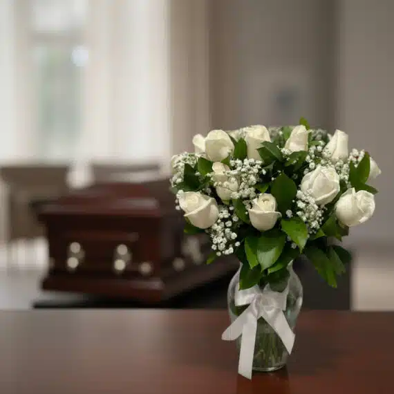 Arreglo floral fúnebre 'Pétalos del Alma' con rosas blancas frescas y follaje verde en jarrón de cristal, sobre mesa de mader