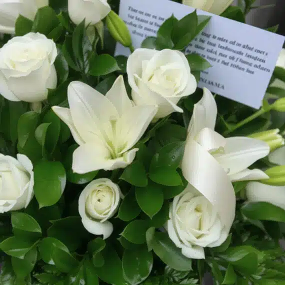 Fotografía macro premium del Arreglo Fúnebre Piedad, con lirios y rosas blancas, follaje verde, rocío sutil y cinta satinada.