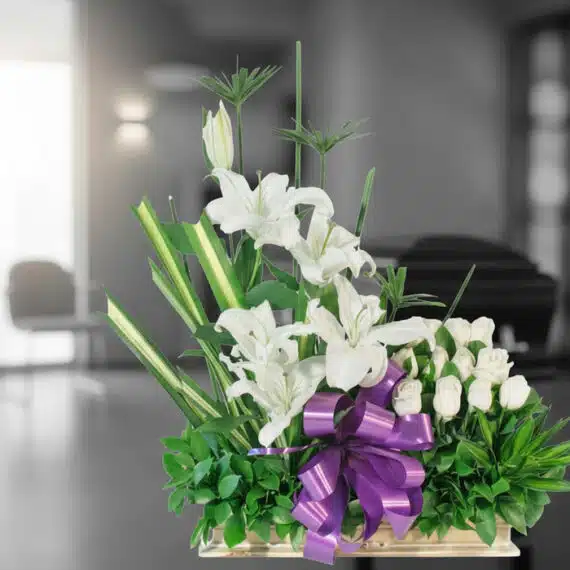Arreglo fúnebre Pureza con lirios y rosas blancas, lazo morado, en sala de velación moderna con ataúd desenfocado al fondo.