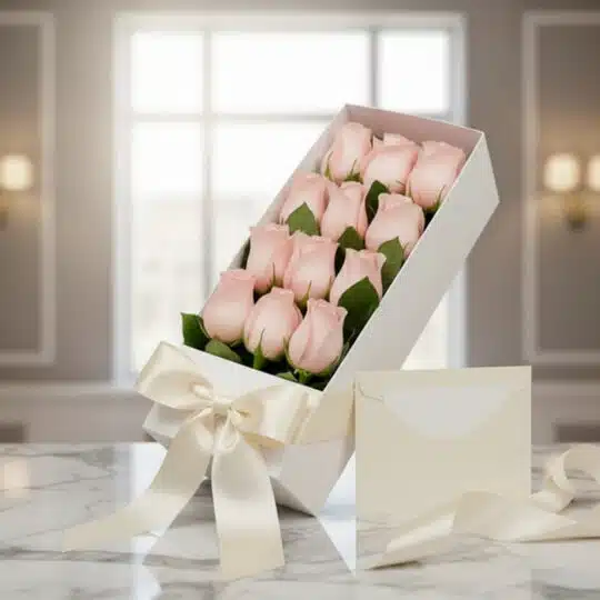 Caja blanca inclinada con doce rosas rosadas frescas, lazo de satén color crema y tarjeta de regalo sobre mármol pulido, con