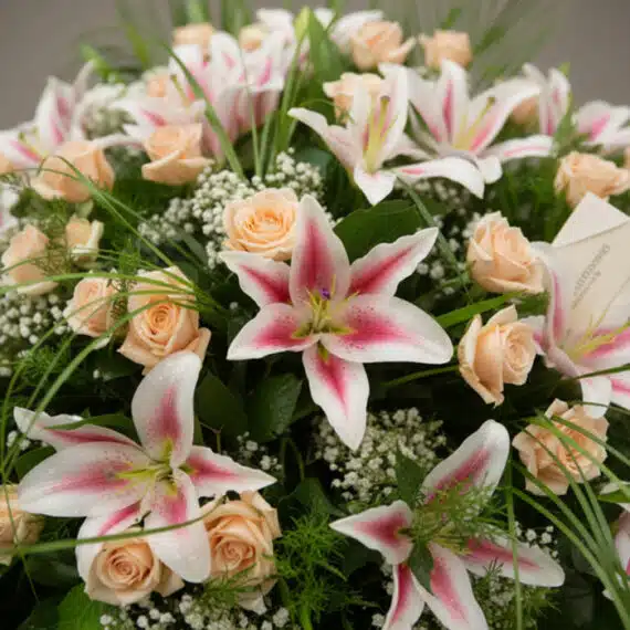 Primer plano de una corona fúnebre 'Luz Eterna' con lirios blancos y rosas melocotón, rocío sutil, cinta satinada y follaje v