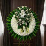 Corona fúnebre grande con rosas blancas, lirios y follaje verde, presentada en una sala de velación premium con luz natural s