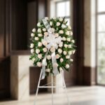 Corona fúnebre Serenidad con rosas blancas y follaje verde, lazo blanco, sobre un trípode en una sala de velación premium con