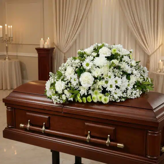 Cubre caja fúnebre Epifania con flores blancas y verdes sobre un ataúd de caoba brillante en una sala de velación de lujo.