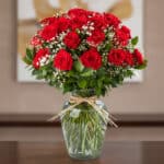 Jarrón de cristal con 24 rosas rojas frescas y follaje verde, atado con un lazo de rafia, sobre una mesa de madera oscura con
